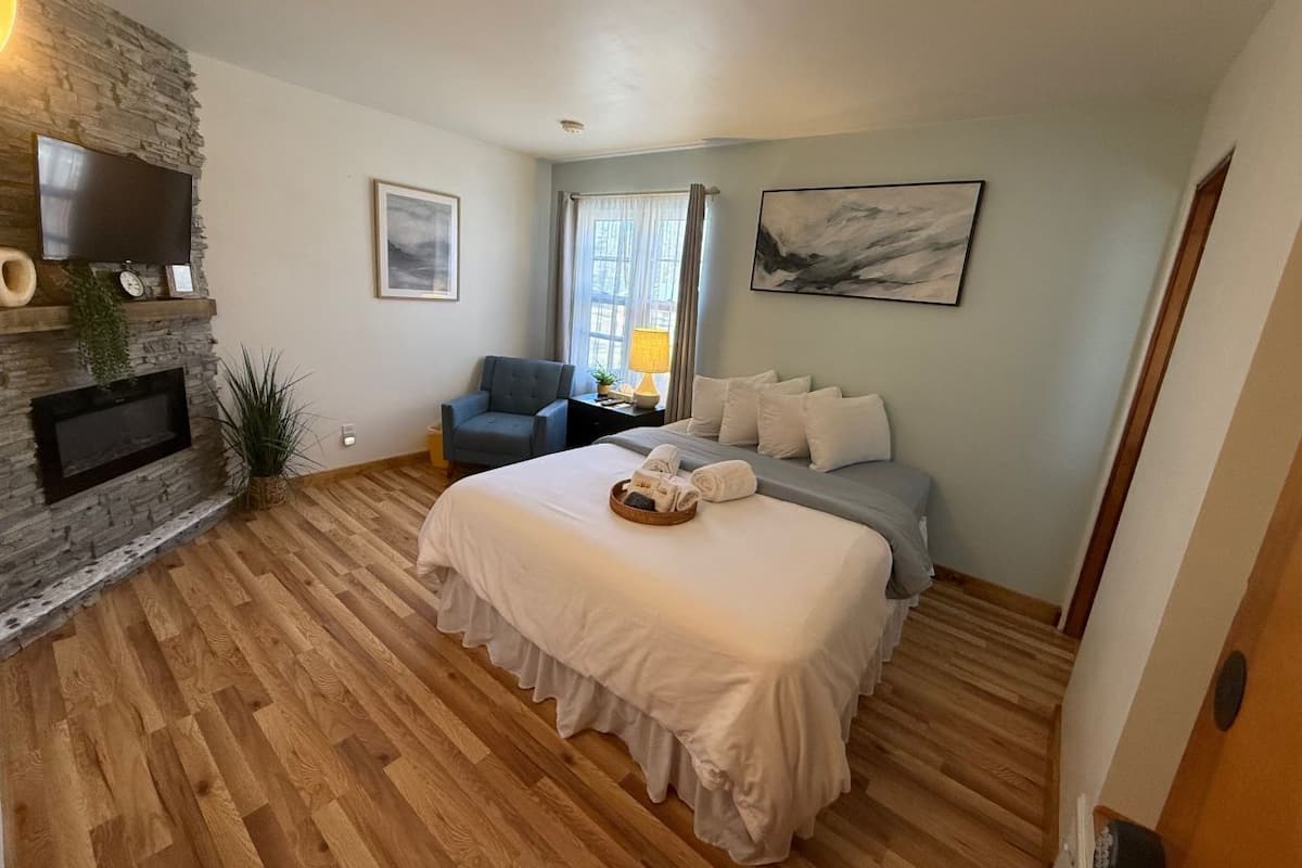 A bedroom with a stone fireplace, a bed with white bedding, a grey armchair, and a large window with curtains.