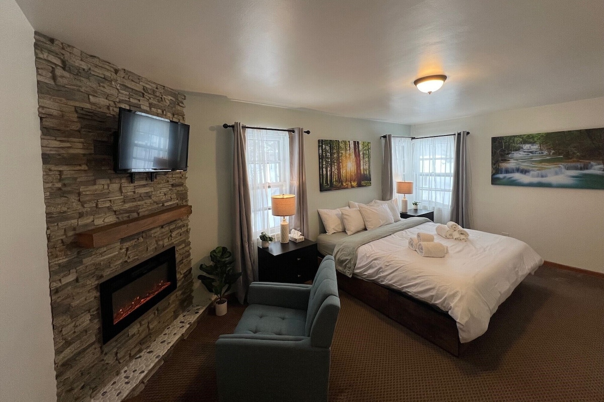 A hotel room with a stone fireplace, a bed with white linens, a TV mounted on the wall, and a painting of a waterfall.
