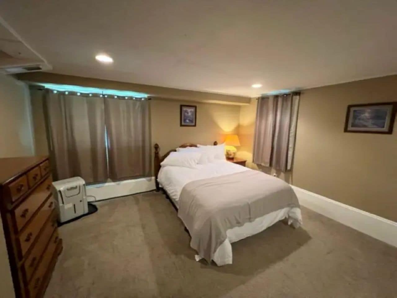 A bedroom with a bed, a dresser, a bedside table with a lamp, and a window with curtains.
