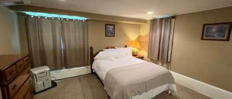 A bedroom with a bed, a dresser, a bedside table with a lamp, and a window with curtains.