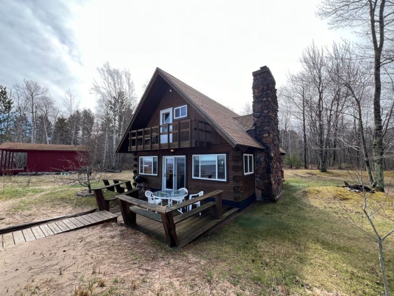 A cabin with a deck, a stone chimney, and a red shed in the background.