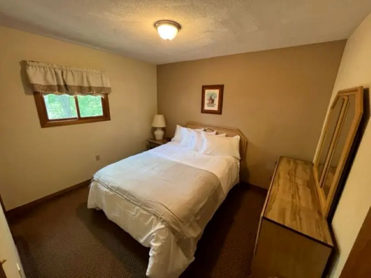 A bedroom with a bed, a nightstand, a dresser, a window with curtains, and a ceiling light.