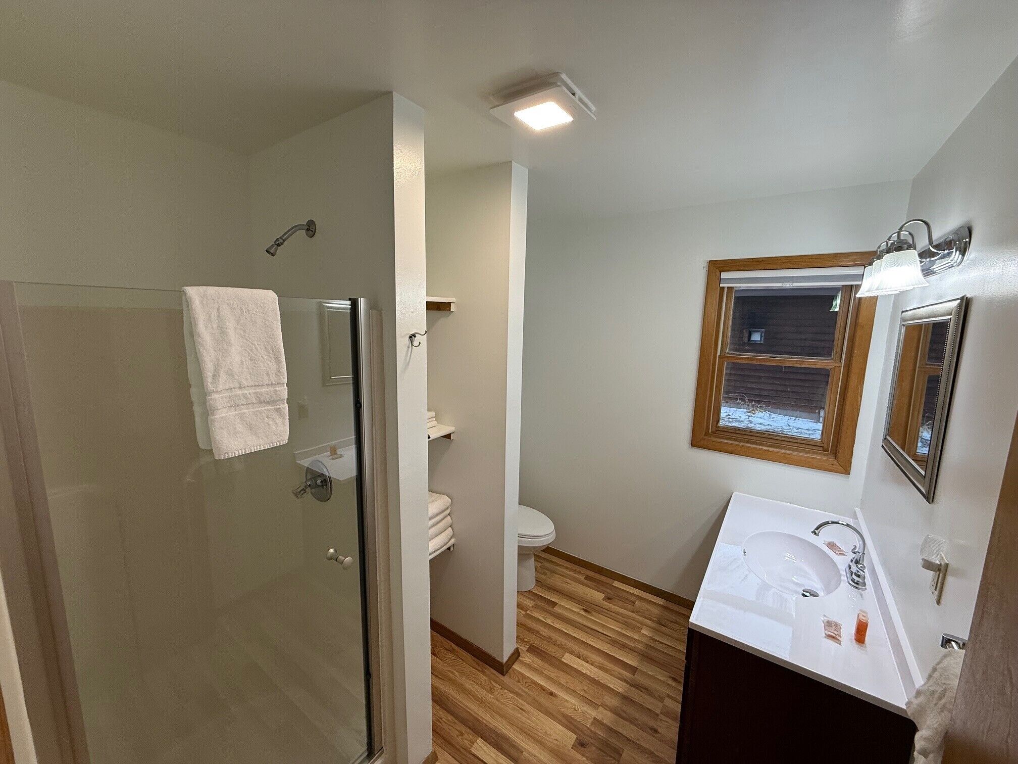 A bathroom with a glass shower enclosure, a toilet, a sink with a mirror, and a window.