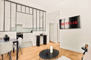 A modern living space with a kitchen, dining area, and a TV mounted on the wall displaying the Netflix logo. - Cœur du Marais – Écrin d'exception pour 4 (Paris)