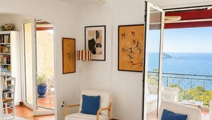 Ein geräumiges Wohnzimmer mit Meerblick, einem Bücherregal, zwei weißen Sesseln mit blauen Kissen und einem Schachtisch.