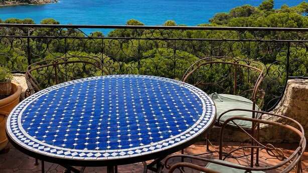 Ein runder Tisch mit einer blau-weißen Mosaikplatte, umgeben von Metallstühlen, auf einer Terrasse mit Blick auf Meer und Grünflächen.