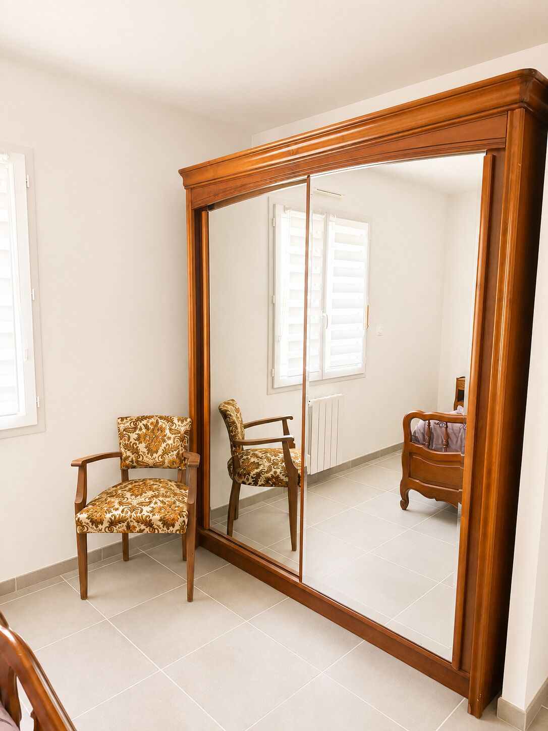 A room with a large mirror, two patterned chairs, a wooden bed, and a radiator.