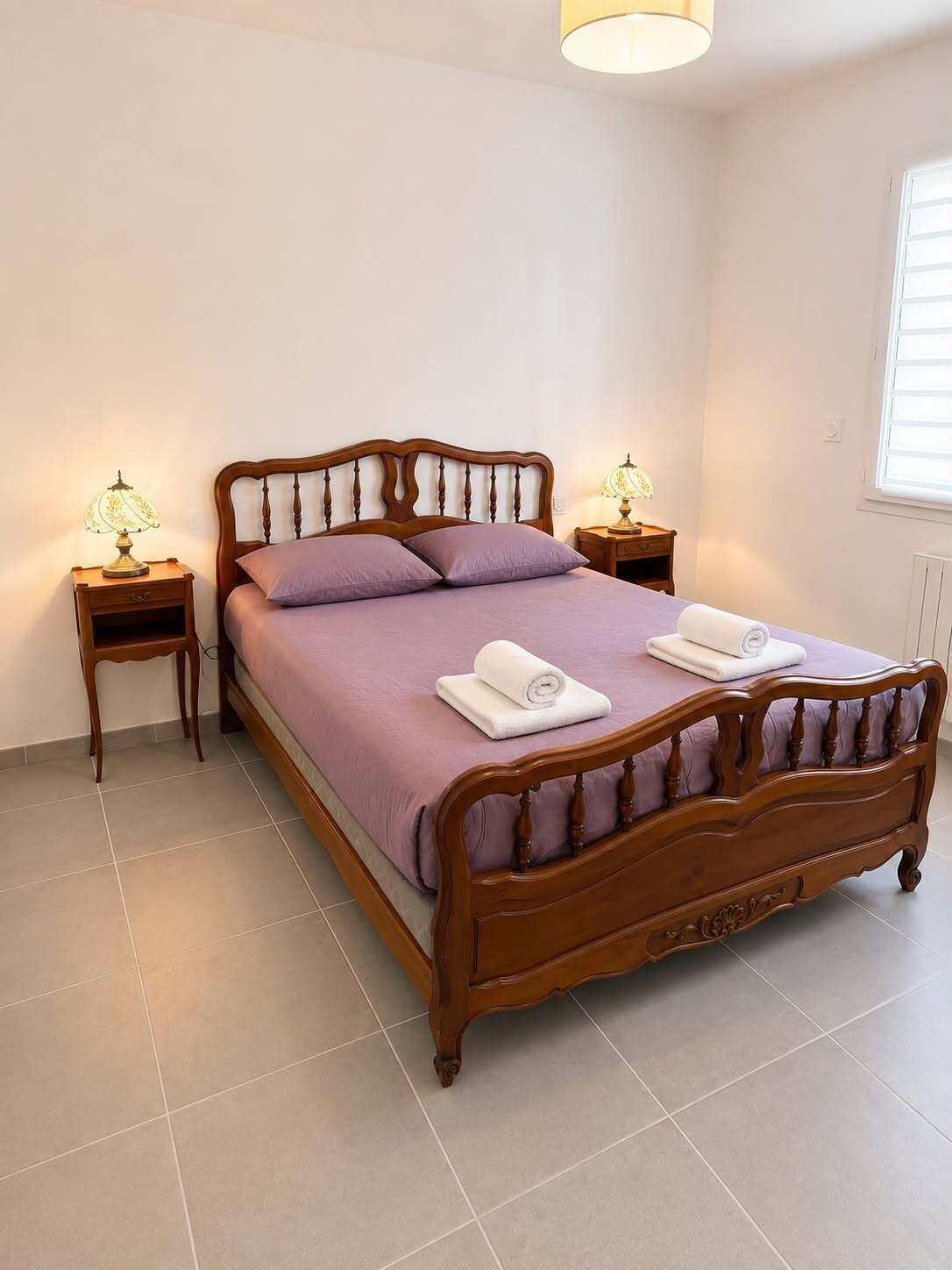 A bedroom with a wooden bed, two bedside tables with lamps, a window with blinds, and a tiled floor.