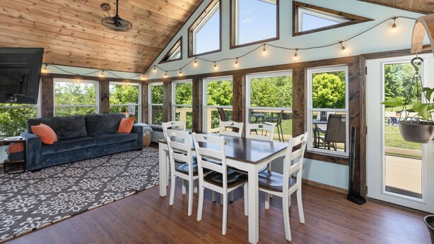 A spacious room with a wooden ceiling, large windows, a dining table with chairs, and a sofa.