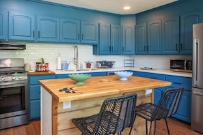 Une cuisine avec des meubles bleus, un îlot central en bois, deux chaises noires, un réfrigérateur en acier inoxydable et un four à micro-ondes.