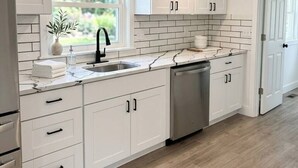 Een moderne keuken met witte kasten, een marmeren aanrechtblad, een ingebouwde vaatwasser en een raam met uitzicht op groen.