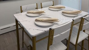 Een eethoek met een witte tafel voor zes personen, gouden stoelen, een stadsgezichtschilderij aan de muur en een raam met zonwering.