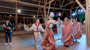 Un grupo de personas con vestimenta tradicional actuando en un espacio abierto con estructura de bambú y suelo de baldosas.