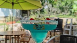 Un cóctel en un vaso con un borde decorativo, sobre una mesa exterior con sillas y una sombrilla al fondo.
