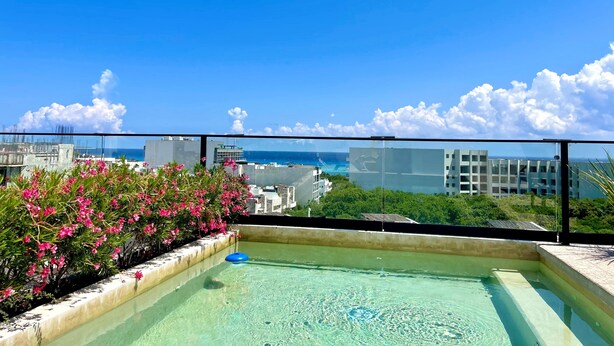 Ein Pool auf dem Dach mit klarem Wasser, umgeben von rosa Blumen und Blick auf das Meer und Gebäude.