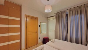 Ein Hotelzimmer mit Holzschrank, Bett mit zusammengerolltem Handtuch, rotem Hocker, Spiegel und einem Fenster mit Vorhängen.