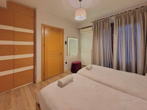 Ein Hotelzimmer mit Holzschrank, Bett mit zusammengerolltem Handtuch, rotem Hocker, Spiegel und einem Fenster mit Vorhängen.