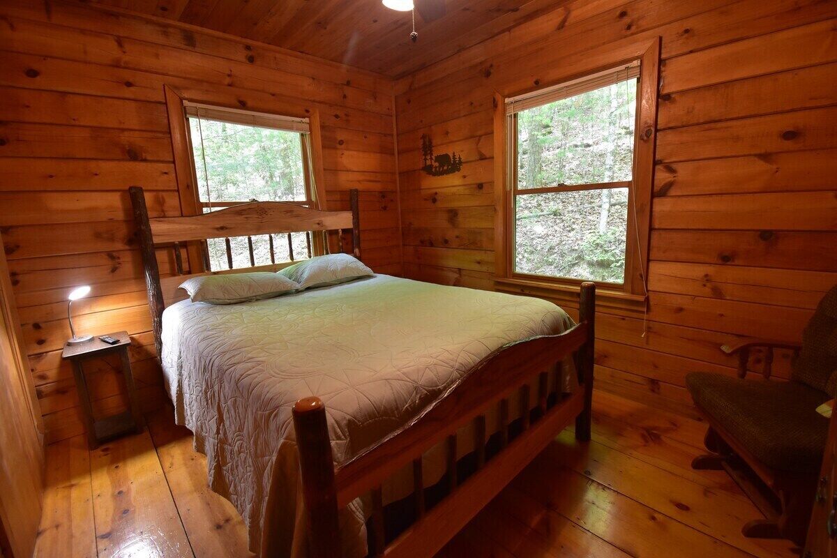 Ein Schlafzimmer in einer Holzhütte mit einem Bett, zwei Fenstern, einem Nachttisch und einem Sessel.