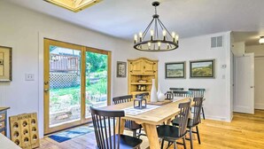 Un coin repas avec une table en bois, des chaises noires, un lustre et une vue sur un jardin, visible à travers une porte vitrée.