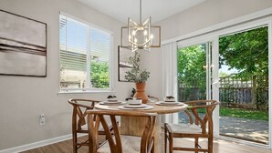 Una sala da pranzo con un tavolo rotondo in legno, quattro sedie, un lampadario e vista su un giardino attraverso una porta scorrevole in vetro.