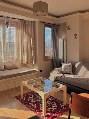 A modern living room with a sofa, a glass coffee table, a patterned rug, and a view of the city through large windows. - Taksim Square1 Bedroom Suite 06 (Istanbul)