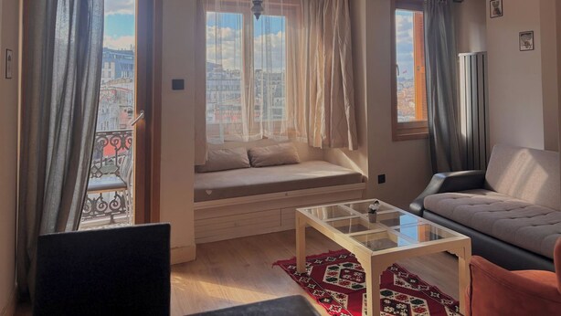 A living room with a sofa, a glass coffee table, a window seat with curtains, and a view of a cityscape. - Taksim Square1 Bedroom Suite 06 (Istanbul)