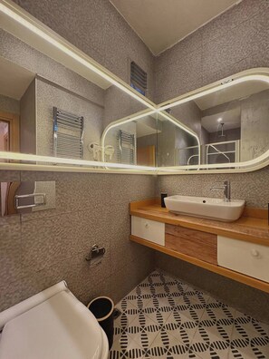A bathroom with a white sink, a mirror with a light strip, a towel rack, and patterned floor tiles. - Taksim Square1 Bedroom Suite 06 (Istanbul)