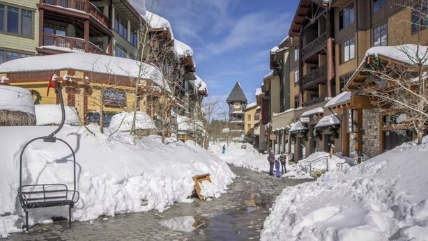 A snow-covered street with buildings, a bench, and a small stream. - Cozy Corner Condo with Shared Pool and Hot Tub, Close to the Lakes & Ski Resort (Mammoth Lakes)