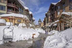 Une rue enneigée bordée d’immeubles, avec un banc et un petit ruisseau.