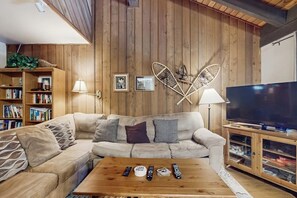 Ein gemütliches Wohnzimmer mit einem Ecksofa, einem Couchtisch mit Fernbedienungen, einem Fernseher und einem Bücherregal.