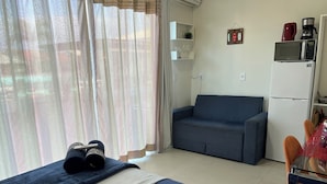 A room with a bed, a sofa, a dining table with chairs, a refrigerator, a microwave, and a coffee maker. - Moradas Dom Goulart  (Palhoça)
