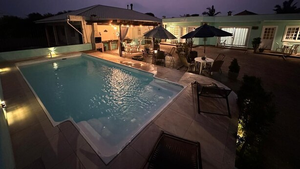 A swimming pool with a covered patio area, outdoor furniture, and a building with windows. - Moradas Dom Goulart  (Palhoça)