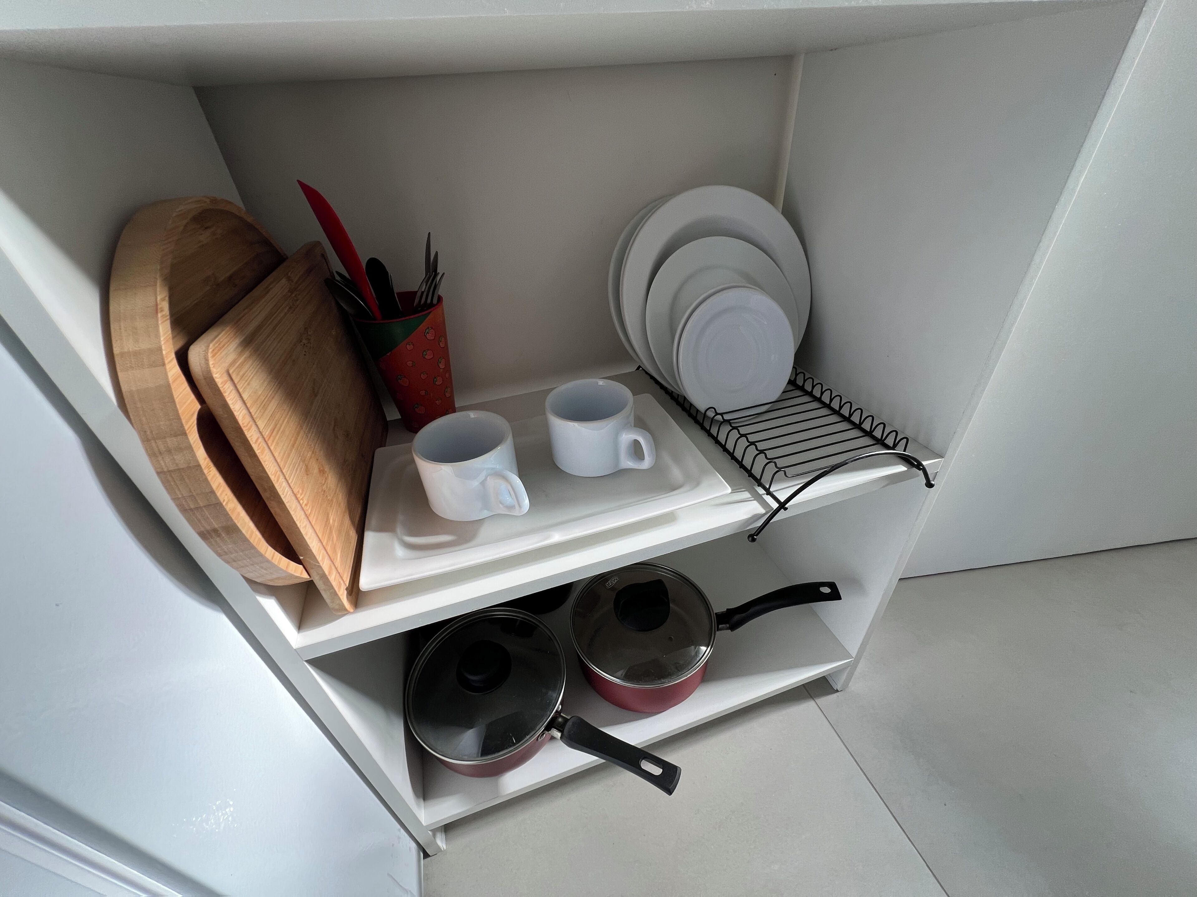 Una zona de cocina con una tabla de cortar de madera, dos tazas blancas, una olla roja y un escurreplatos.