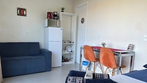 A compact living space with a sofa, a small dining table with chairs, a kitchenette, and a bed with pillows. - Moradas Dom Goulart  (Palhoça)