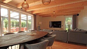 Una sala de estar moderna con techo de madera, ventanas amplias, una mesa de comedor y un sofá gris.