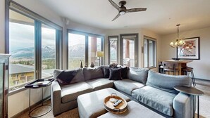 Ein modernes Wohnzimmer mit einer Eckcouch, einem Couchtisch, einem Essbereich mit Tisch und Stühlen und einem großen Fenster mit Blick auf die Berge.