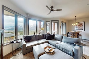 Ein modernes Wohnzimmer mit einer Eckcouch, einem Couchtisch, einem Essbereich mit Tisch und Stühlen und einem großen Fenster mit Blick auf die Berge.