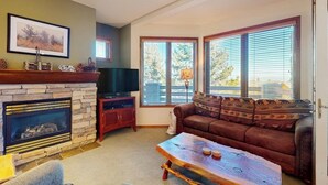 Una sala de estar con chimenea de piedra, televisor, sofá de cuero y ventanas amplias.
