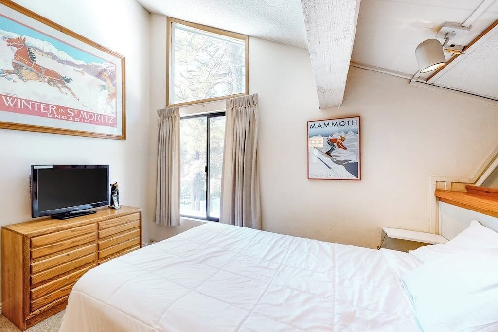 Une chambre à coucher avec un lit, un téléviseur posé sur une commode en bois, une fenêtre avec des rideaux et deux tableaux encadrés accrochés au mur.