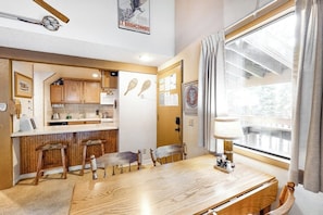 A dining area with a wooden table, four chairs, a lamp, and a window with curtains. - Cozy Studio with a Loft, Shared Swimming Pool, Hot Tub, and Sauna (Mammoth Lakes)