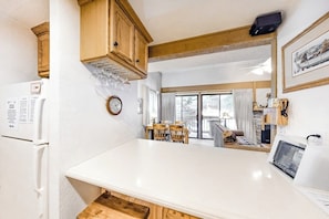 A kitchen with a white countertop, wooden cabinets, a refrigerator, a clock, and a dining area with chairs and a table. - Cozy Studio with a Loft, Shared Swimming Pool, Hot Tub, and Sauna (Mammoth Lakes)
