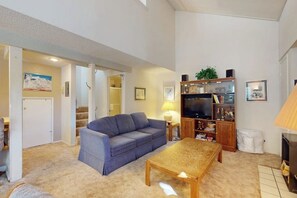 A living room with a sofa, a coffee table, a TV, a bookshelf, and a lamp. - Mountain-view, two-level studio with decks, pool & hot tub - close to skiing (Mammoth Lakes)