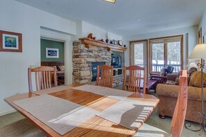 A dining area with a wooden table and chairs, a stone fireplace, and a sofa in a well-lit room with large windows. - Mountain Getaway with a Shared Outdoor Pool, Hot Tub and Nature Views (Mammoth Lakes)