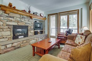 A stone fireplace with a TV, a wooden table, and a sofa in a living room with a view of trees. - Mountain Getaway with a Shared Outdoor Pool, Hot Tub and Nature Views (Mammoth Lakes)