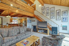 Ein Wohnzimmer mit einer Couch, einem Couchtisch, einem Fernseher und Lautsprechern. Eine Treppe führt zu einer oberen Ebene.