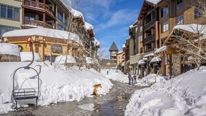 Una calle cubierta de nieve con edificios, un banco y un telesilla.