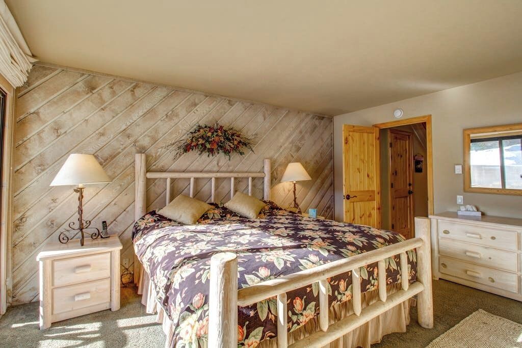 Une chambre à coucher avec une tête de lit en bois, une couvre-lit à motifs floraux, deux tables de chevet avec des lampes, une commode en bois et une porte.