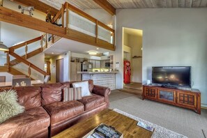 Ein großzügiger Wohnbereich mit Ledersofa, Couchtisch aus Holz und Fernseher auf einer Holzkommode. Eine Treppe führt zu einer oberen Ebene.