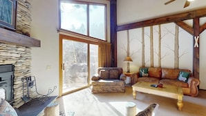A living room with a stone fireplace, a sofa set, a coffee table, and a large window. - Townhome Near Skiing with Fireplace & Shared Pool, Hot Tub Access, Dog-Friendly (Mammoth Lakes)