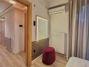 A hotel room with a wooden floor, a red ottoman, a radiator, an air conditioning unit, and a window with curtains. - Taksim Square1 Bedroom Suite 03 (Istanbul)